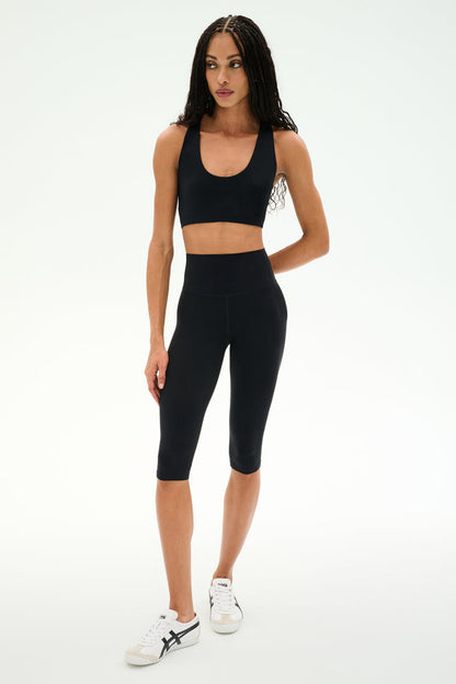 A woman stands against a plain white background wearing the SPLITS59 Sara Airweight Bra in black, paired with high-waisted black bike shorts and white sneakers—an ideal outfit for Pilates or hot yoga.