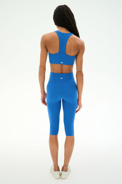 A person is shown from the back, wearing the SPLITS59 Sara Airweight Bra in Classic Blue along with matching high-waisted leggings and light-colored shoes, set against a plain white background.