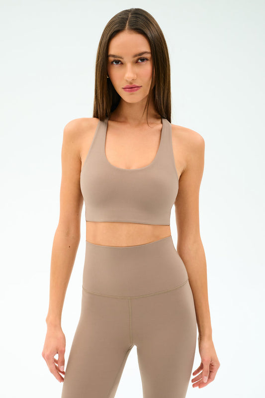 A woman models the SPLITS59 Sara Airweight Bra in Latte with matching high-waisted leggings against a plain white background—ideal for hot yoga or Pilates.