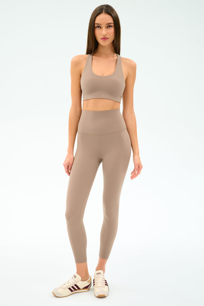 A woman stands against a plain background wearing the SPLITS59 Sara Airweight Bra in Latte, matching high-waisted leggings ideal for Pilates or hot yoga, and beige sneakers with maroon stripes.