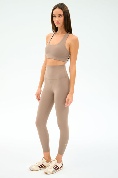 A woman in a SPLITS59 Sara Airweight Bra in Latte stands against a plain white background, ready for Pilates or hot yoga.