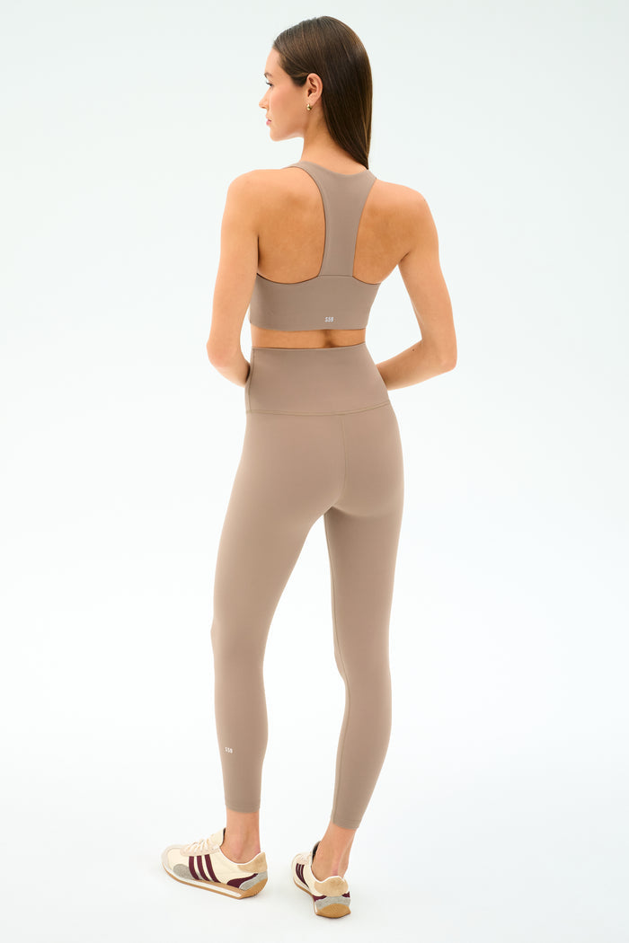 A woman stands facing away in the SPLITS59 Sara Airweight Bra - Latte and matching high-waisted leggings—ideal for Pilates or hot yoga—with beige sneakers, against a plain light background.