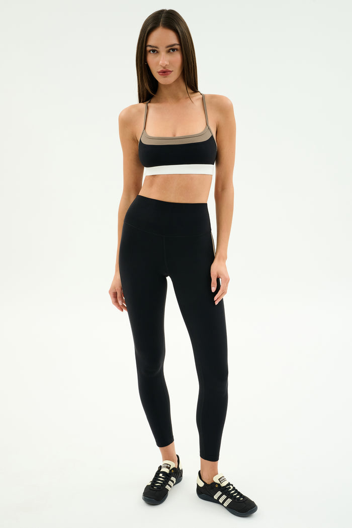A woman stands against a plain background, wearing the SPLITS59 Scottie Airweight Bra in Black/Lentil, paired with high-waisted black leggings and athletic shoes.