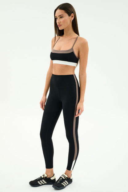 Woman stands in studio wearing the SPLITS59 Scottie Airweight Bra in Black/Lentil and matching high-waisted leggings with beige side stripes, paired with black Adidas sneakers.