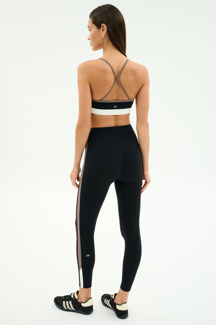 A woman stands on a plain background, facing away, wearing the SPLITS59 Scottie Airweight Bra in Black/Lentil, paired with black leggings featuring a side stripe and black sneakers.