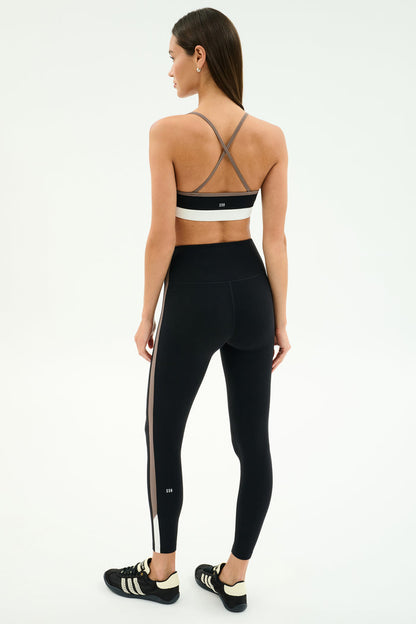 A woman stands on a plain background, facing away, wearing the SPLITS59 Scottie Airweight Bra in Black/Lentil, paired with black leggings featuring a side stripe and black sneakers.