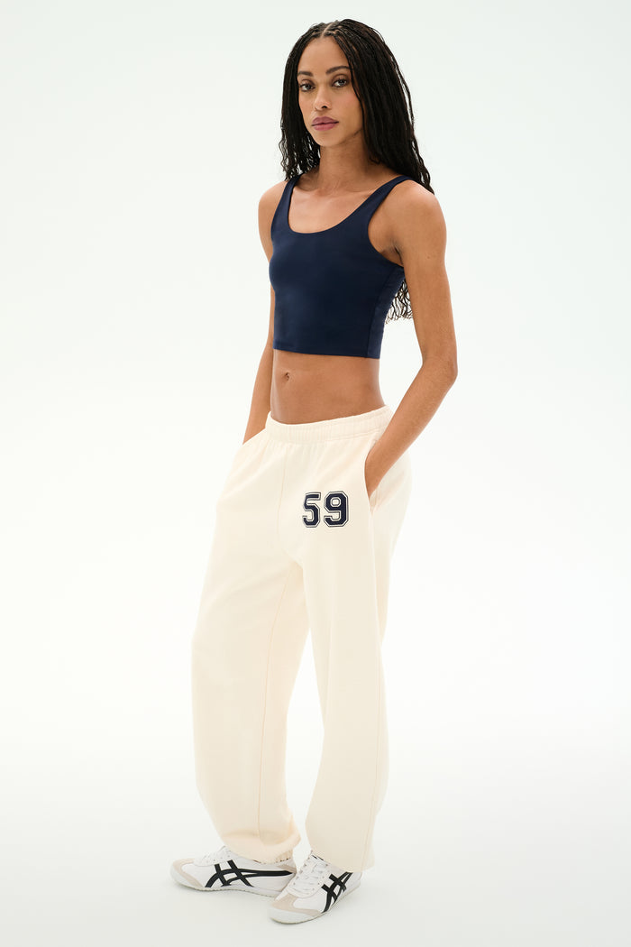 A woman stands against a white background wearing a navy crop top, white sneakers with black stripes, and SPLITS59 Serena French Terry Oversized Sweatpant in Creme/59—crafted from soft cotton and featuring "59" on the hip.