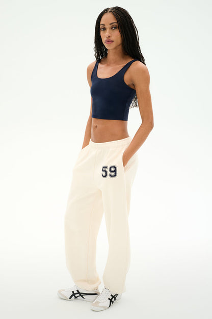 A woman stands against a white background wearing a navy crop top, white sneakers with black stripes, and SPLITS59 Serena French Terry Oversized Sweatpant in Creme/59—crafted from soft cotton and featuring "59" on the hip.