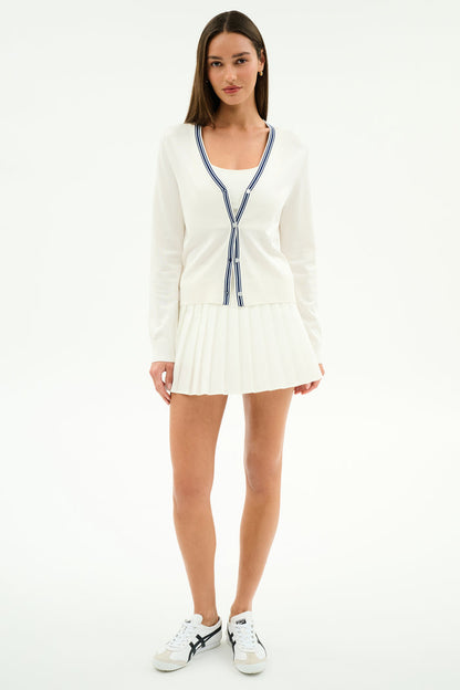 A woman faces forward against a plain background, wearing the SPLITS59 Shira Woven Pleated Skort in white and a white V-neck cardigan with blue trim, paired with white sneakers.