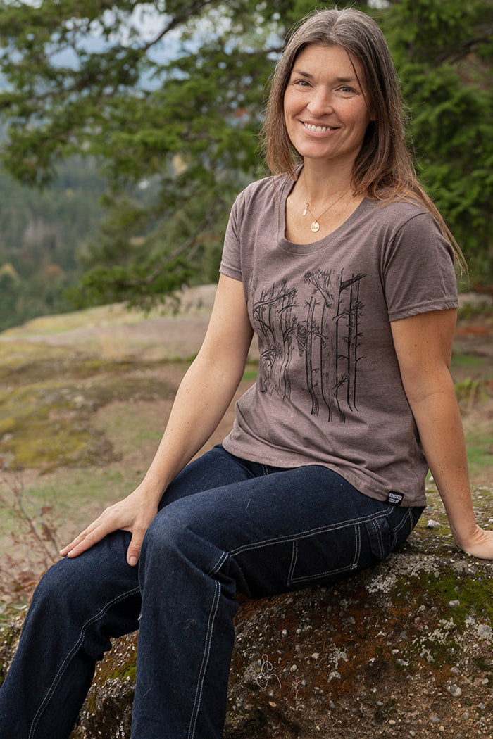 Woman sitting on a rock with a scenic background of trees and mountains porint on her tee of trees and an owl