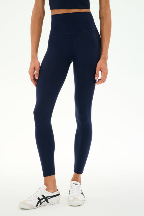 A person wearing SPLITS59 Sprint High Waist Rigor 7/8 leggings in Indigo, crafted from soft Rigor fabric, paired with a matching crop top and white sneakers, stands against a plain white background.
