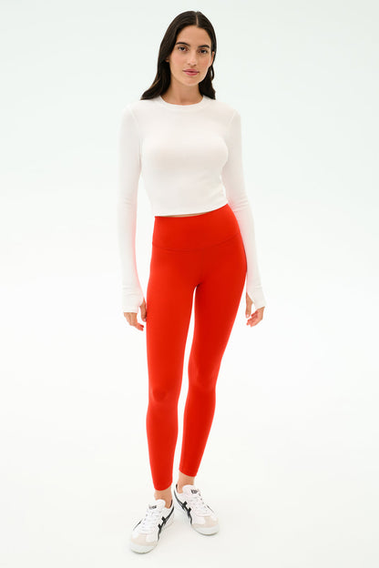 A person stands facing forward in a white long-sleeve top, white sneakers, and SPLITS59 Sprint High Waist Rigor 7/8 leggings in Lotus Red, against a plain white background.