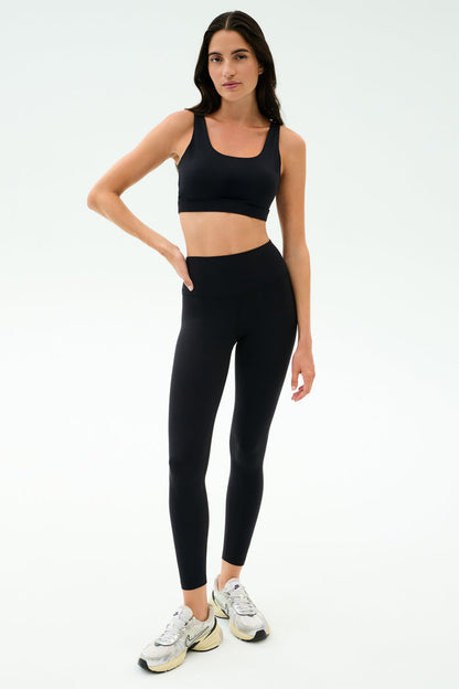 A woman stands on a white background wearing the black SPLITS59 Sprint Rigor Bra, paired with black high-waisted leggings and white sneakers.