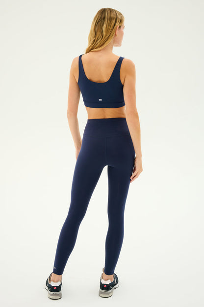 A woman with long blonde hair stands facing away, wearing a SPLITS59 Sprint Rigor Bra in Indigo, high-waisted leggings, and black sneakers against a plain light background. Her outfit exemplifies multi-sport performance, ideal for diverse gym workouts.