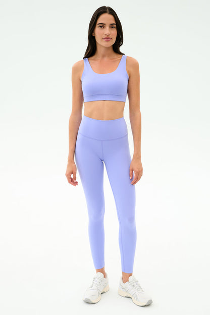 A woman stands facing forward in the SPLITS59 Sprint Rigor Bra - Purple Haze and matching leggings with white athletic shoes, designed for multi-sport performance, against a plain white background.
