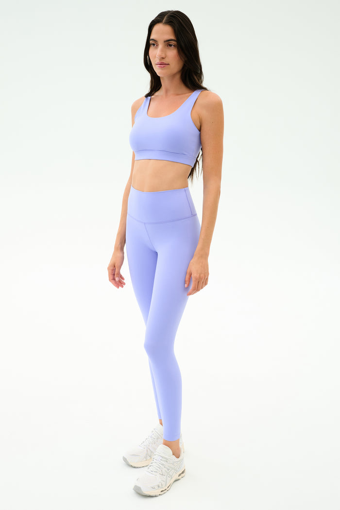 A woman wears the SPLITS59 Sprint Rigor Bra in Purple Haze and matching leggings, styled with white sneakers against a plain light background.