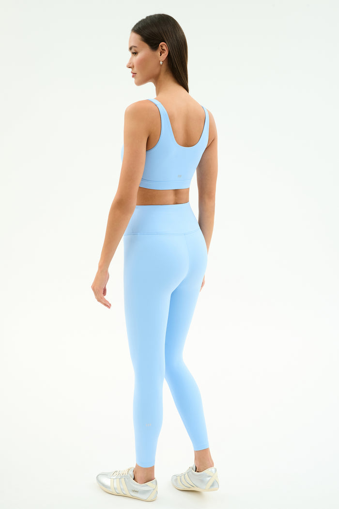 A woman faces away, wearing the SPLITS59 Sprint Rigor Bra in Skylight and matching leggings with white athletic shoes, ready for gym workouts or multi-sport performance against a plain background.