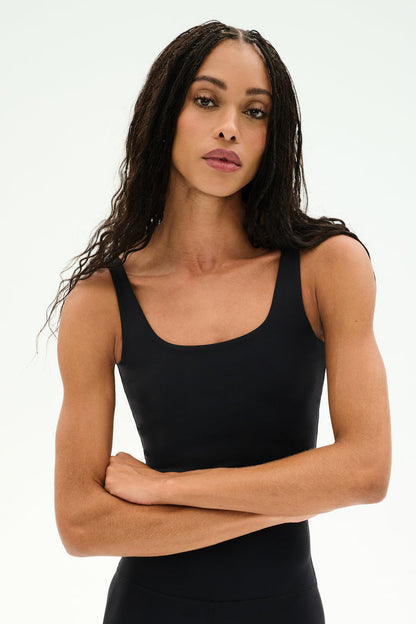 A person with long braided hair stands against a plain light background, arms crossed, wearing the SPLITS59 Sprint Rigor Bralette in black—an athletic crop tank designed for multisport performance.