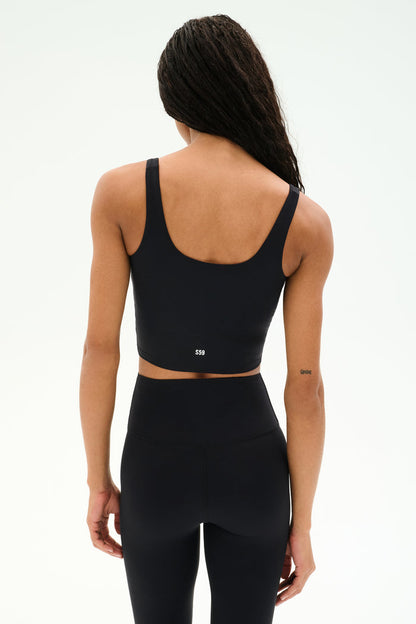 A person stands with their back to the camera, highlighting the SPLITS59 Sprint Rigor Bralette in black paired with high-waisted black leggings, set against a plain light background.