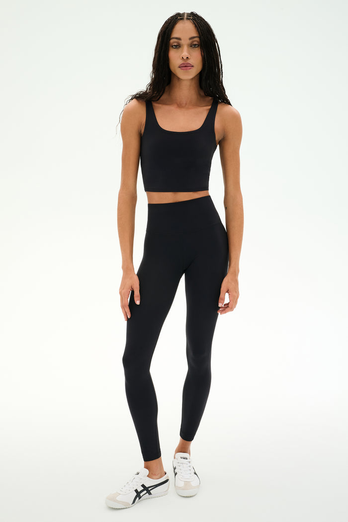 A person stands against a white background wearing the SPLITS59 Sprint Rigor Bralette in black, paired with black leggings and white sneakers with black accents, demonstrating versatile multisport performance and stylish sports bra support.