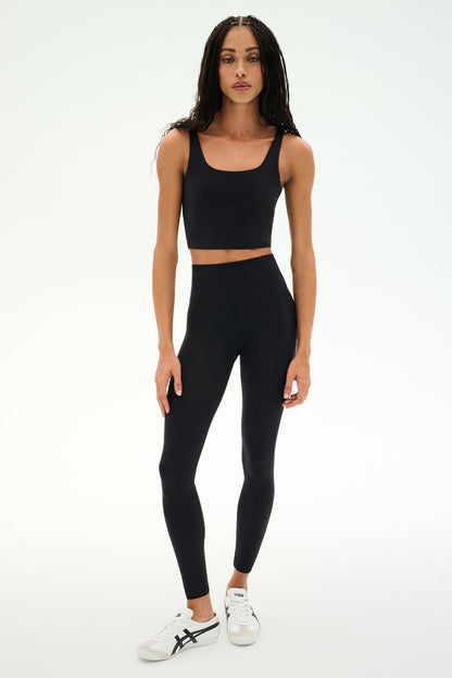 A person stands against a white background wearing the SPLITS59 Sprint Rigor Bralette in black, paired with black leggings and white sneakers with black accents, demonstrating versatile multisport performance and stylish sports bra support.