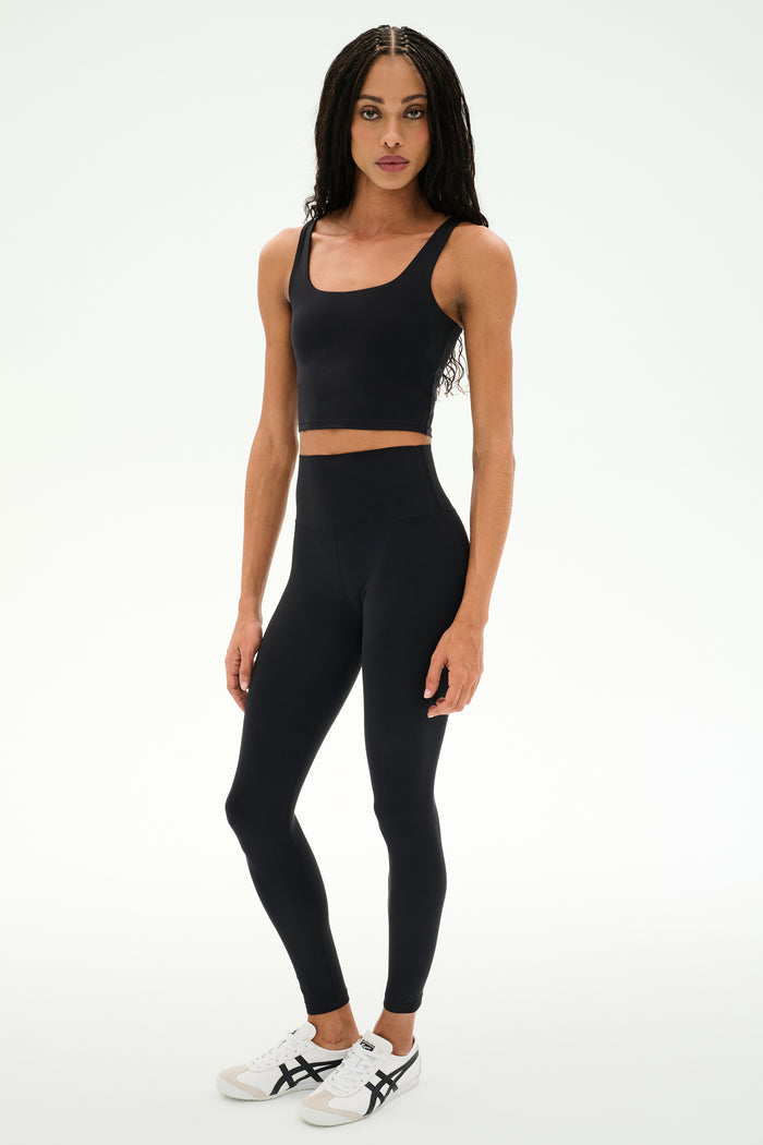 A person wears the SPLITS59 Sprint Rigor Bralette in black paired with high-waisted black leggings and white sneakers, standing against a plain light background—ideal for multi-sport performance.