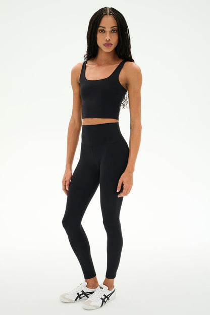 A person wears the SPLITS59 Sprint Rigor Bralette in black paired with high-waisted black leggings and white sneakers, standing against a plain light background—ideal for multi-sport performance.