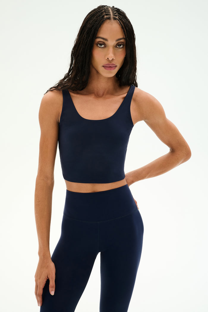 A woman with long braids wears the SPLITS59 Sprint Rigor Bralette in Indigo, paired with matching high-waisted leggings, standing against a plain white background—the perfect look for multi-sport performance.