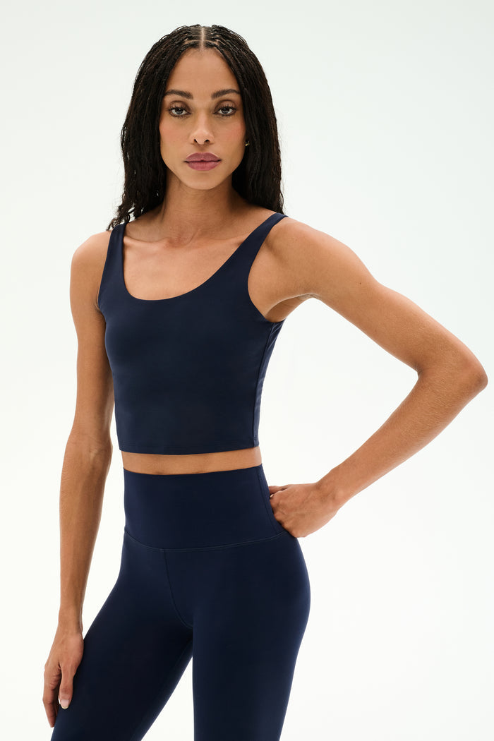 A woman stands against a plain background with one hand on her hip, wearing the SPLITS59 Sprint Rigor Bralette in Indigo and matching high-waisted leggings, highlighting multi-sport performance style and supportive sports bra design.
