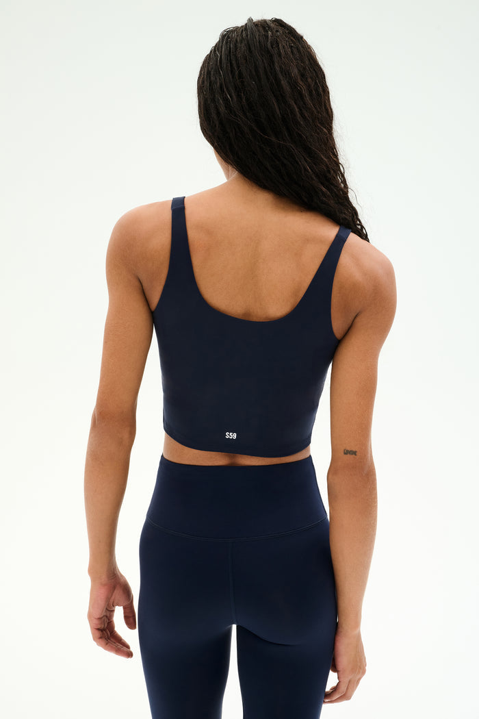 A woman with long braided hair, seen from the back, wears the SPLITS59 Sprint Rigor Bralette in Indigo—crafted for multi-sport performance—paired with matching high-waisted leggings against a plain white background.