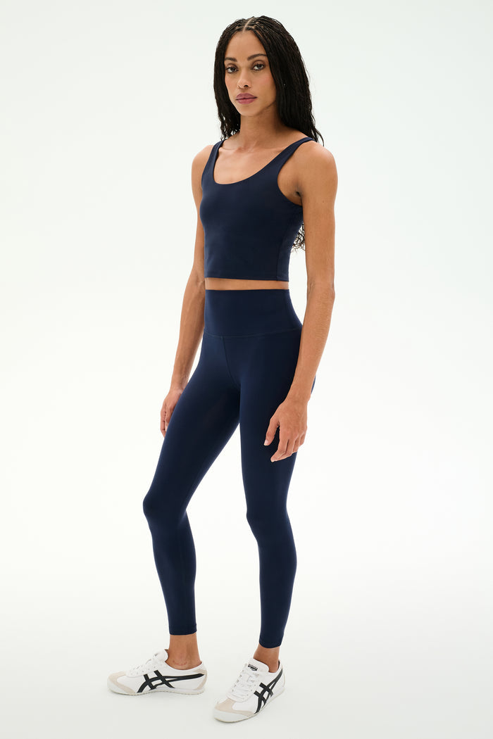 A woman stands against a plain white background wearing the SPLITS59 Sprint Rigor Bralette in Indigo with matching high-waisted leggings and white sneakers.