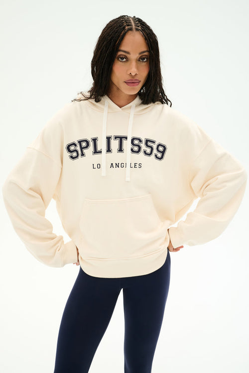 A person wearing the SPLITS59 Stella French Terry Oversized Hoodie in Creme/Splits59 Los Angeles, featuring soft cotton fabric, and navy leggings stands with hands on hips against a plain white background.