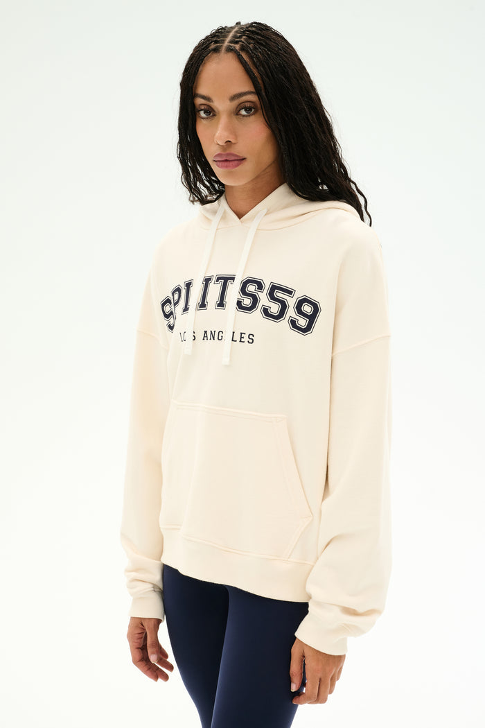 A woman with long braids wears the SPLITS59 Stella French Terry Oversized Hoodie in Creme/Splits59 Los Angeles—a soft 100% cotton sweatshirt featuring signature "SPLITS59 LOS ANGELES" text—paired with dark leggings, standing against a plain white background.