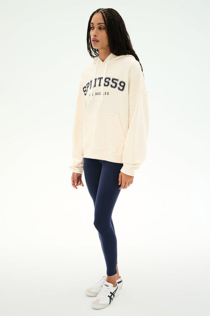 A woman stands against a plain white background wearing the SPLITS59 Stella French Terry Oversized Hoodie in Creme/Splits59 Los Angeles, paired with navy leggings and white sneakers.