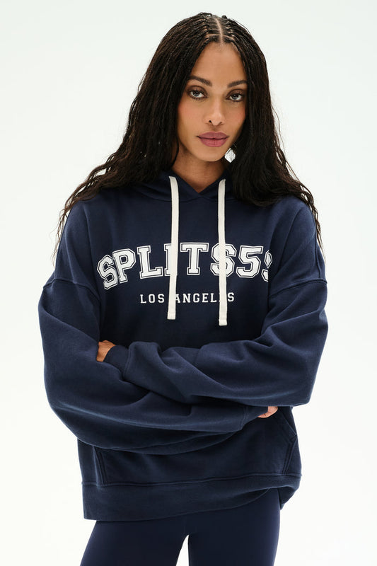 A woman stands with arms crossed against a plain background, wearing the SPLITS59 Stella French Terry Oversized Hoodie in Indigo—a soft 100% cotton piece featuring "SPLITS59 LOS ANGELES" printed on the front.