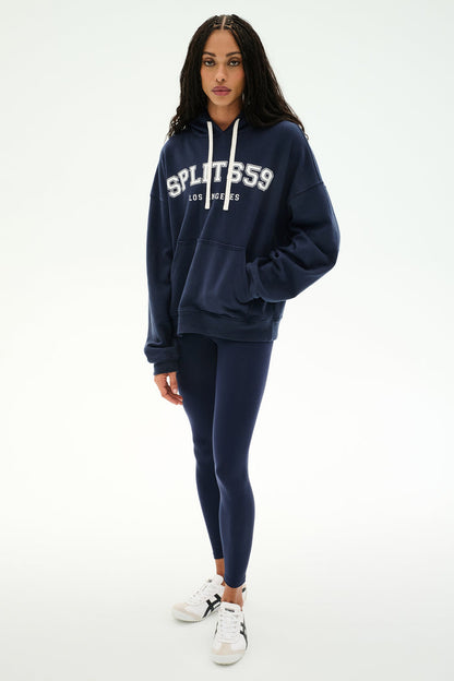 A woman stands facing forward, wearing the SPLITS59 Stella French Terry Oversized Hoodie in Indigo/Splits59 Los Angeles, crafted from soft cotton fabric. She pairs it with navy leggings and white sneakers against a plain white background.