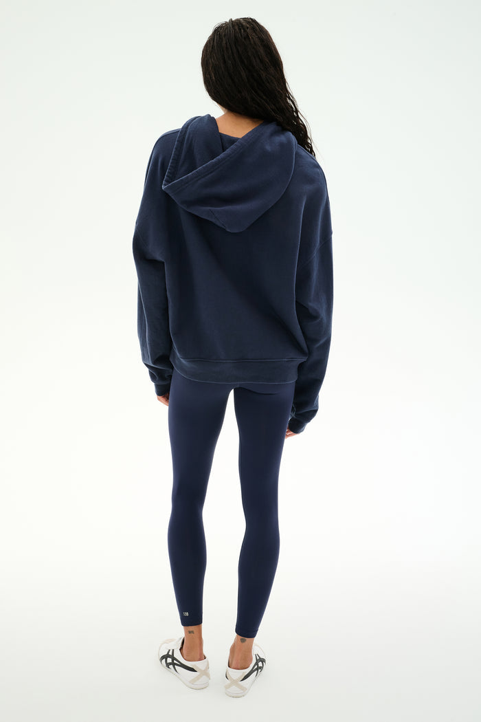 Standing with back to camera, the person is wearing the SPLITS59 Stella French Terry Oversized Hoodie in Indigo, paired with navy leggings and white sneakers against a plain white background.