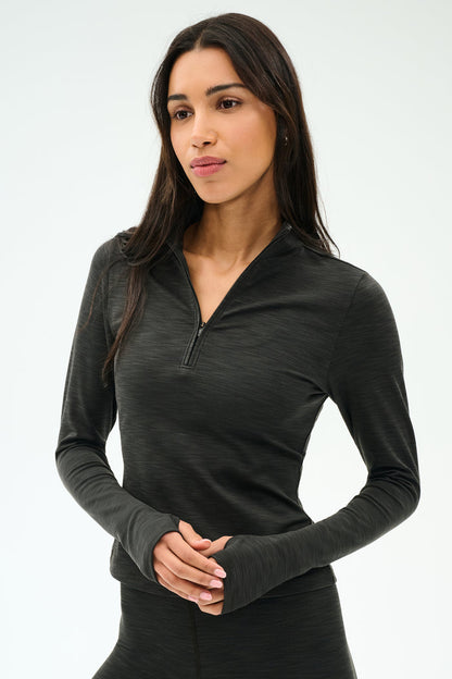 A woman with long dark hair wears the SPLITS59 Stevie Active Rib Half Zip Waist Length top in Heather Graphite, standing against a plain light background.