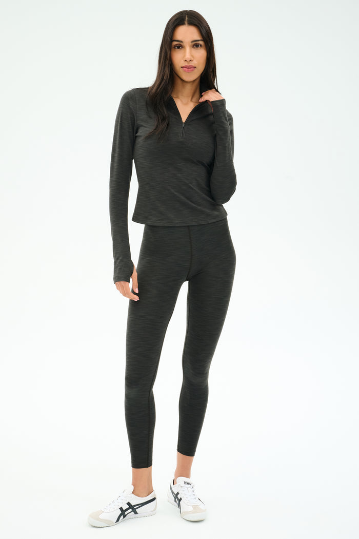 A woman stands in a studio wearing the SPLITS59 Stevie Active Rib Half Zip Waist Length top in Heather Graphite, paired with matching leggings and white sneakers, with a neutral expression.