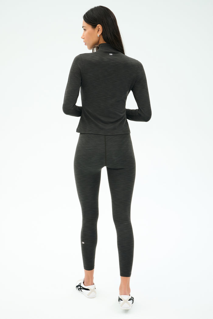 A woman faces away, wearing the SPLITS59 Stevie Active Rib Half Zip Waist Length in Heather Graphite with matching leggings and white athletic shoes, against a plain white background.