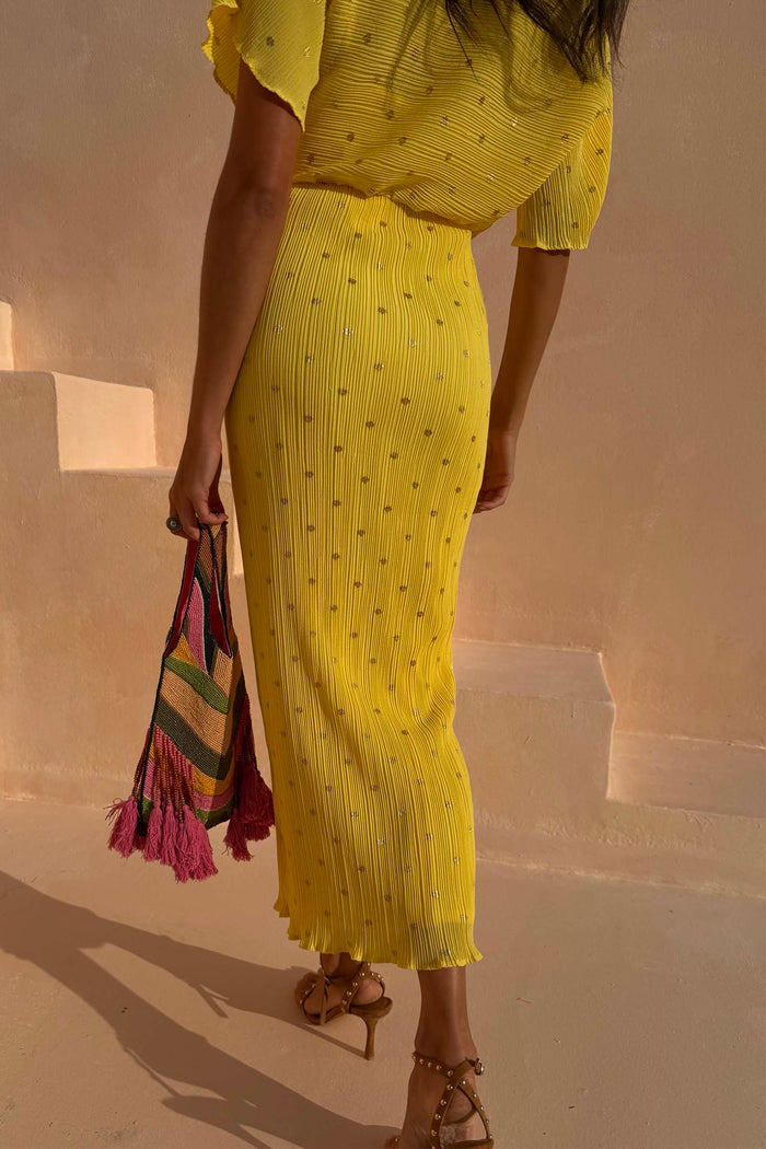 Sunshine Tilly Dress in yellow plissé with gold flecks and lettuce hem, held with a colourful beaded bag.