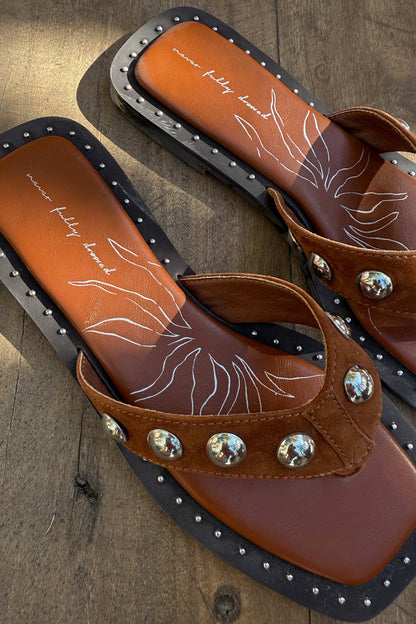 Tan leather stud sandals with silver studs, a square toe and suede straps on a wooden background.