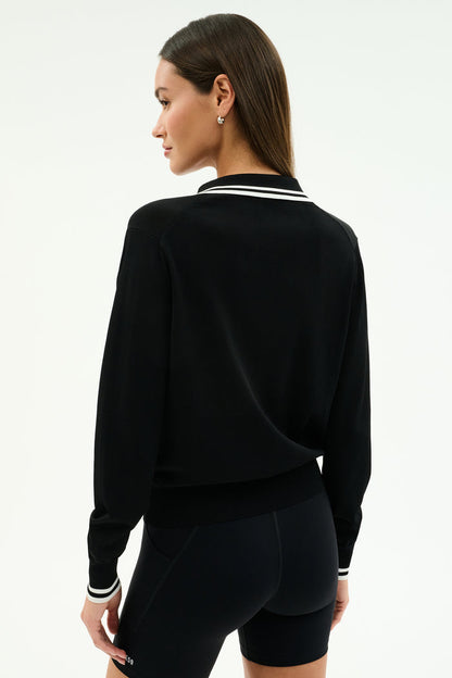 A woman stands with her back to the camera in the SPLITS59 Taylor Knit Polo in Black/White, a soft rayon-nylon blend long-sleeve top featuring white trim, styled with black fitted shorts against a plain light background.
