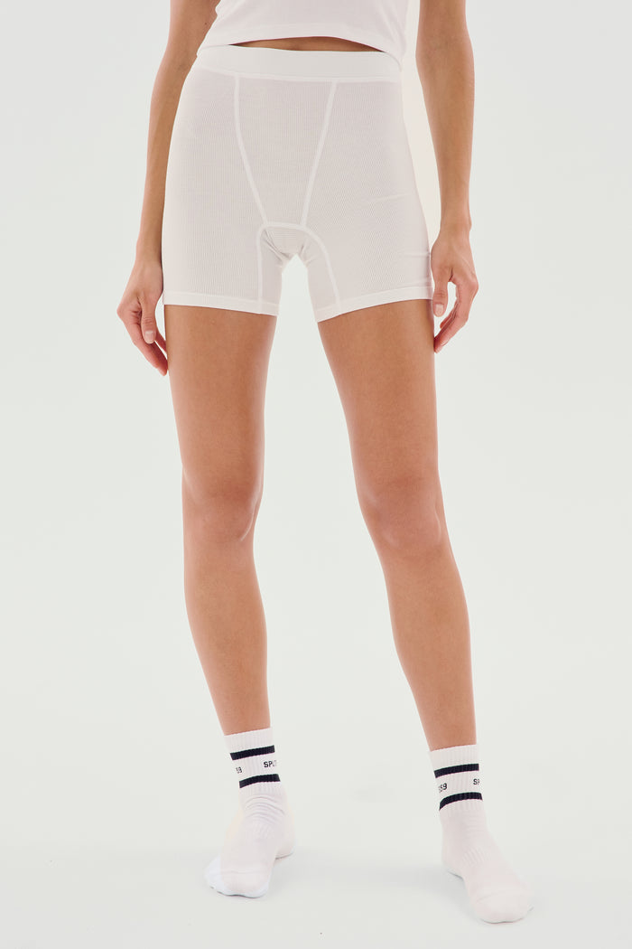Person wearing white, mid-thigh Theo Rib Short - White from SPLITS59, a white top, and white socks with black stripes, standing against a plain white background. A perfect blend of tailored comfort and classic design.