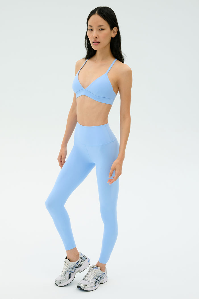 A person wears the SPLITS59 Theo Rigor Bra in Skylight, styled with matching high-waisted leggings and white athletic shoes against a plain light background.