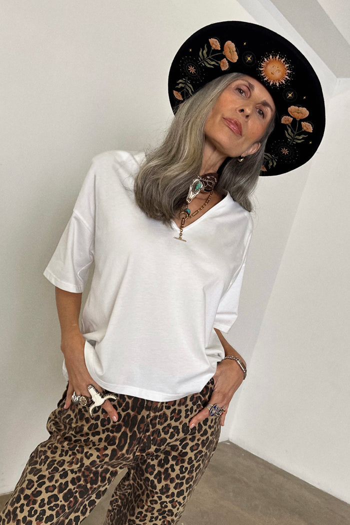 White cotton T-shirt with shoulder pads and a deep V-neck, styled with leopard print trousers and a patterned hat.
