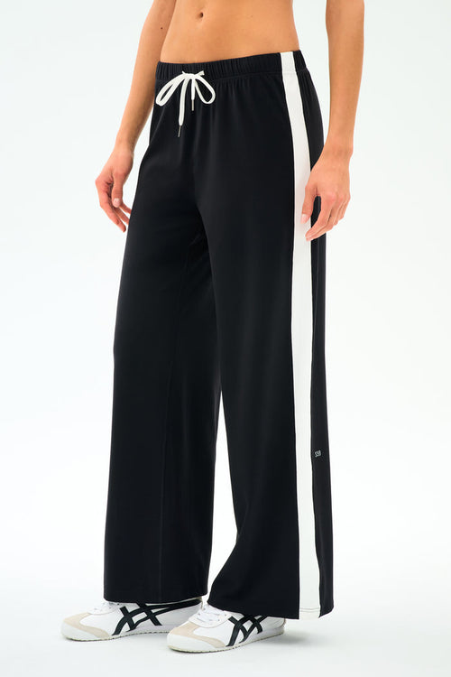 A person models the SPLITS59 Willow Airweight Wide Leg pants in Black/White, featuring buttery-soft fabric, wide legs, white side stripes, and a white drawstring, paired with white sneakers against a plain background.
