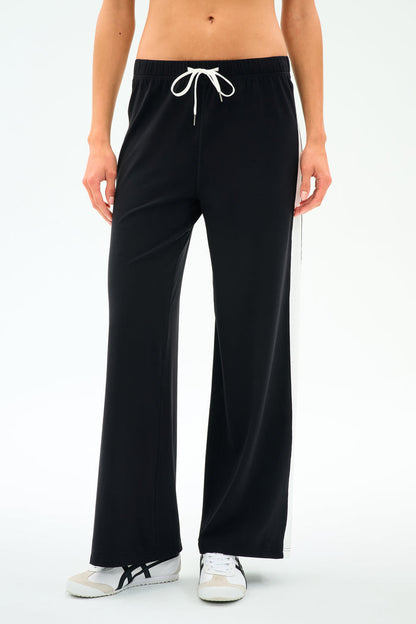 A person wearing SPLITS59's Willow Airweight Wide Leg pants in black with white side stripes and white sneakers, standing against a plain white background.