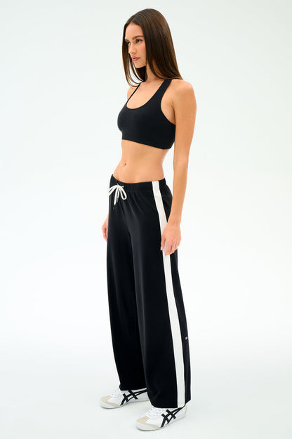 A woman stands against a white background wearing SPLITS59’s Willow Airweight Wide Leg - Black/White pants and a black sports bra with white sneakers—perfect for Pilates.