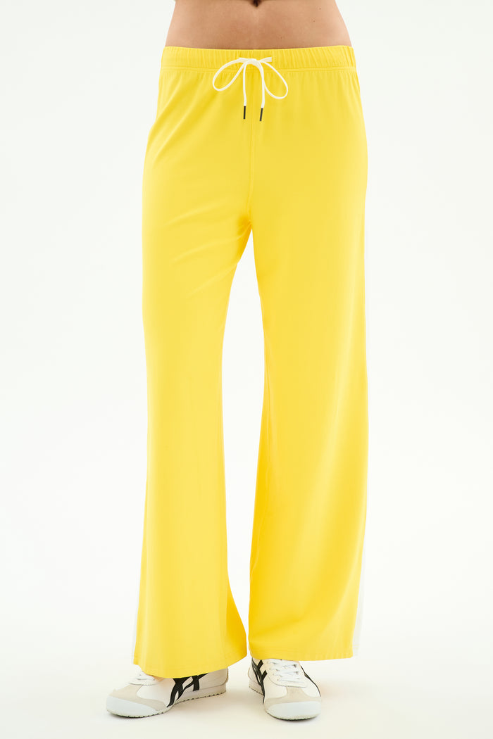 A person wears SPLITS59 Willow Airweight Wide Leg pants in Bright Yellow/White, crafted from a soft nylon-spandex blend, paired with white sneakers against a plain white background.